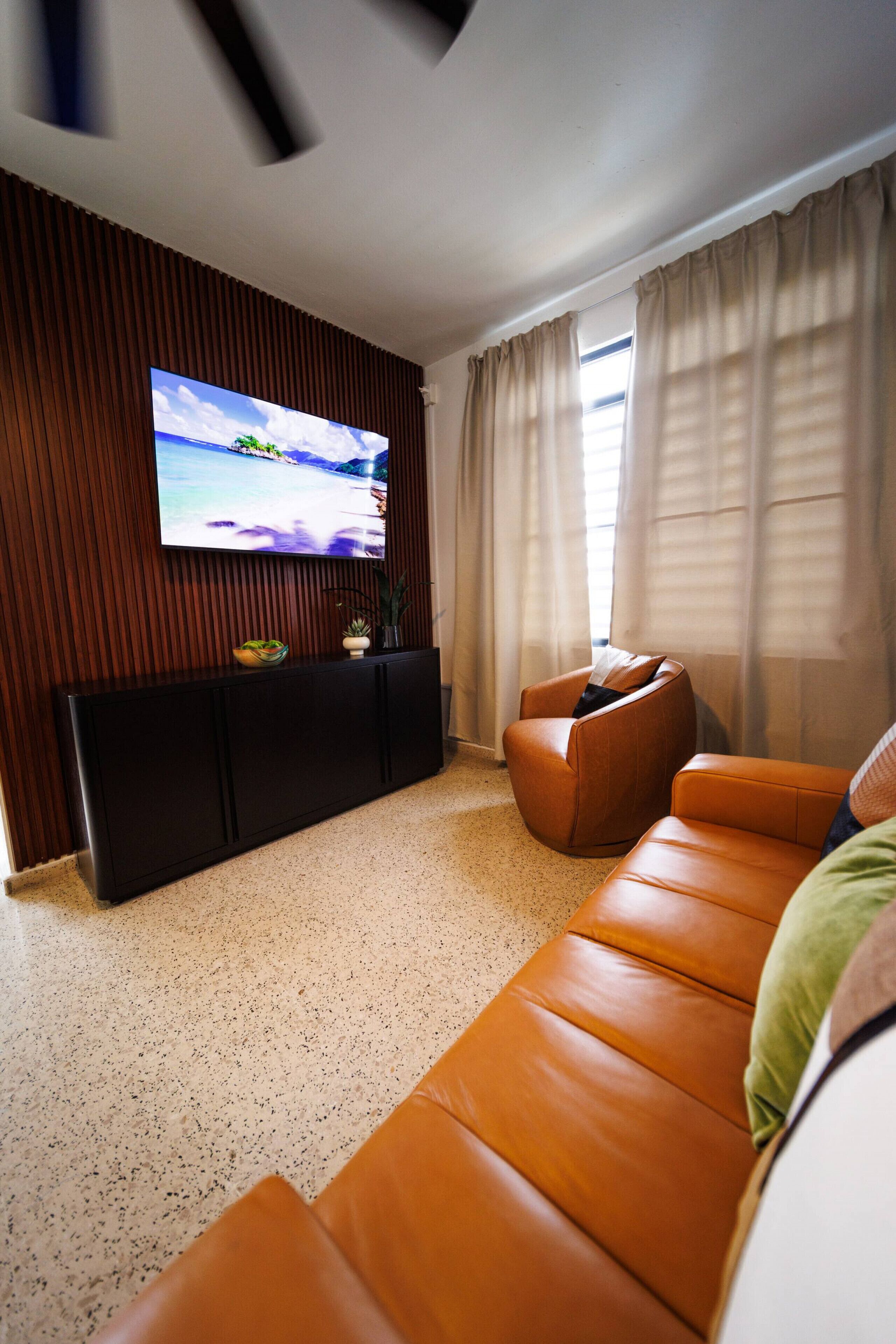 En moderne stue med et fladskærms-tv monteret på en væg af træpaneler, en brun lædersofa og et vindue med lette gardiner.