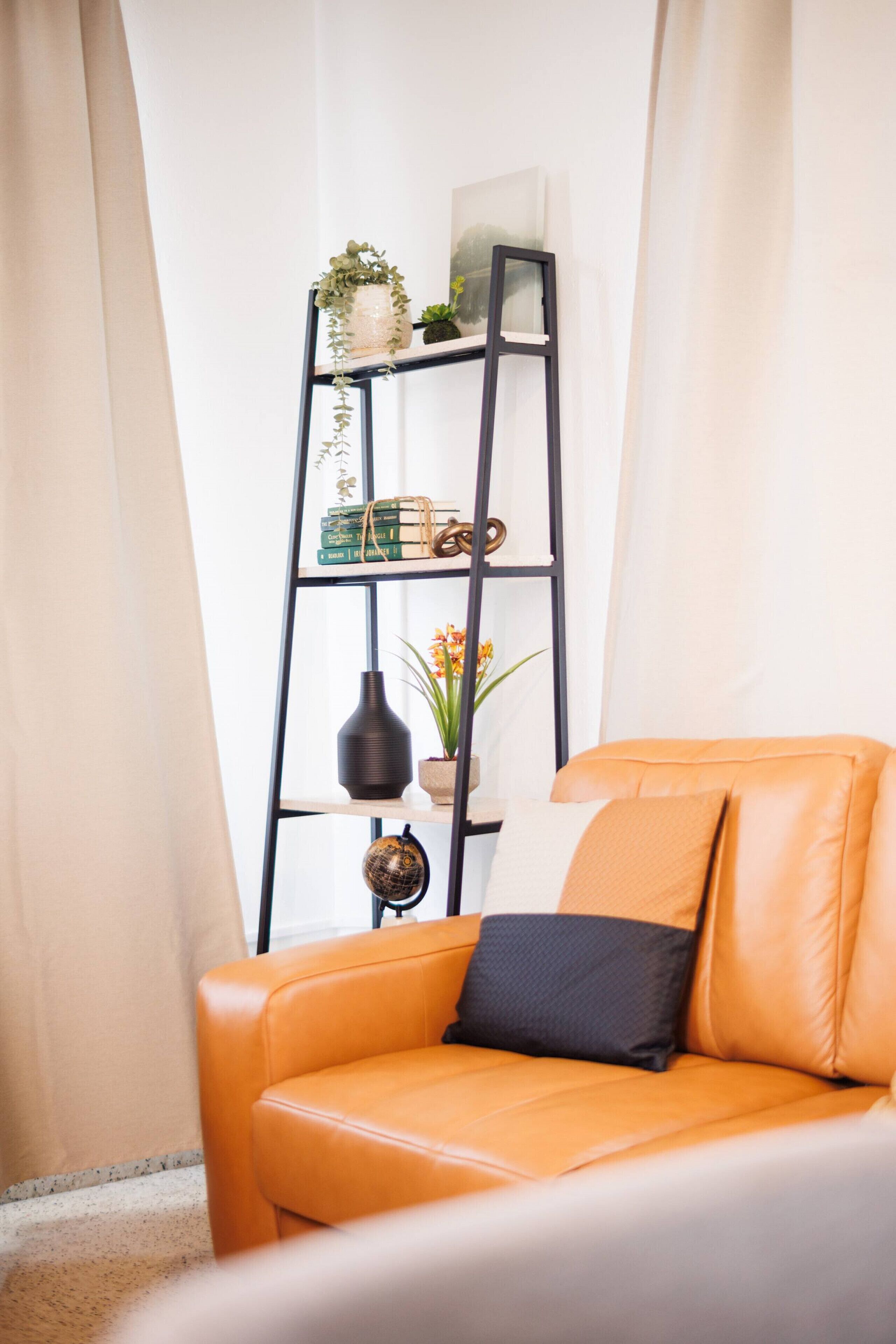 Una sala de estar moderna con un sofá de cuero, una estantería de metal negro con objetos decorativos y una ventana con cortinas traslúcidas.