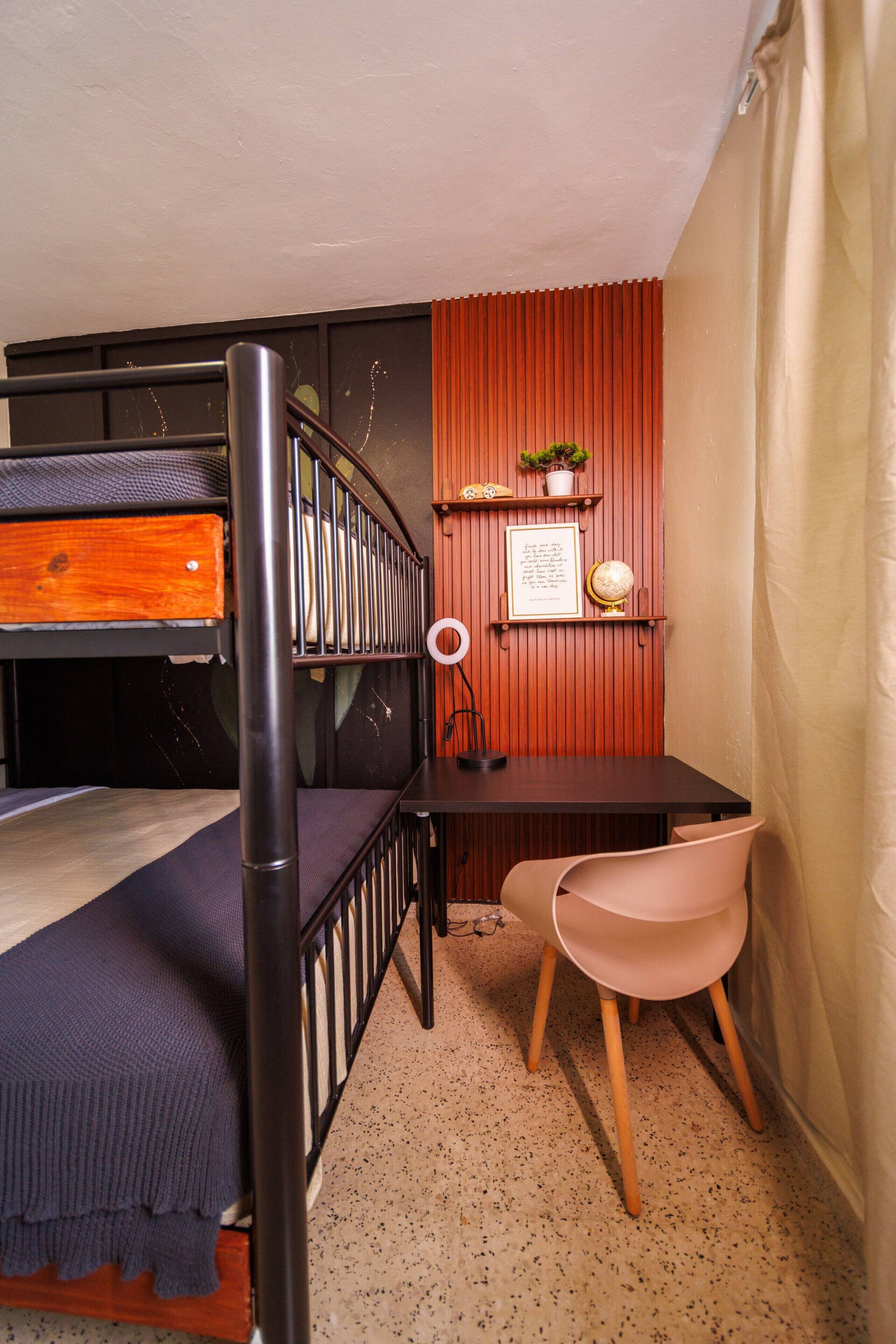 Ein Studentenwohnheimzimmer mit Etagenbett, Schreibtisch, Globus, gerahmtem Zitat, Stuhl und kleinem Regal mit Pflanze.