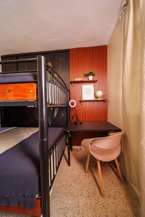 A dormitory room with a bunk bed, a desk with a globe and a framed quote, a chair, and a small shelf with a plant.