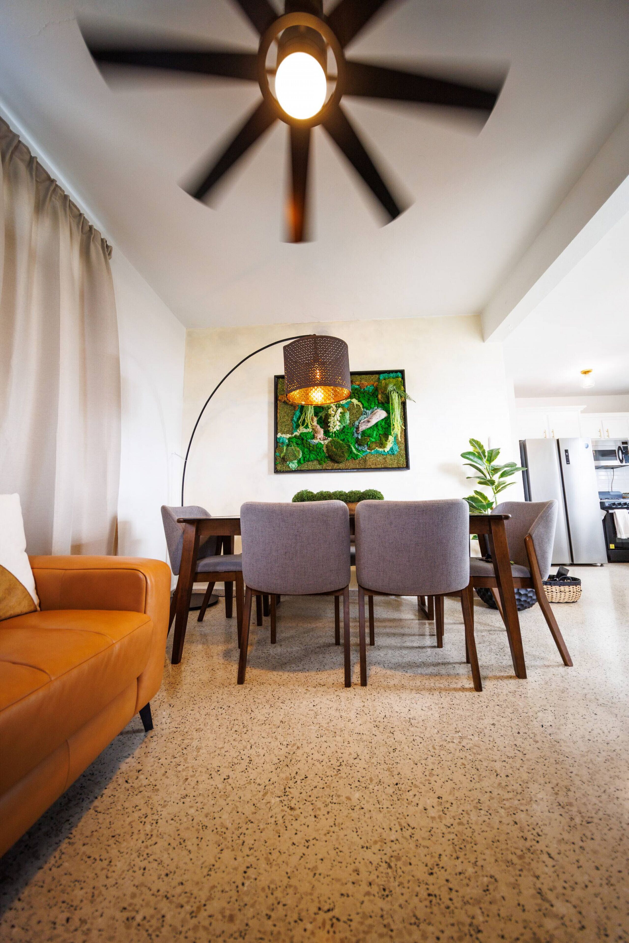 Ein modernes Wohnzimmer mit Deckenventilator, einem Esstisch mit Stühlen, einer Couch und einer Küche im Hintergrund.