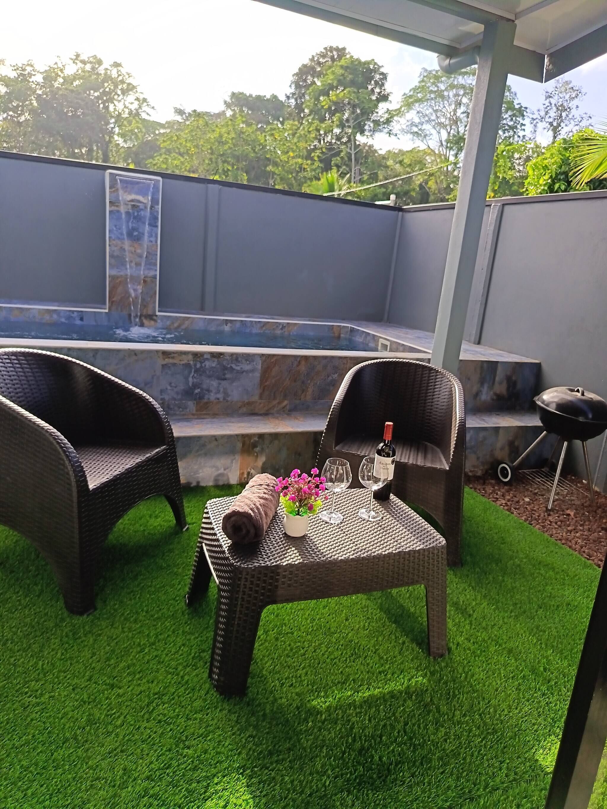 Une terrasse extérieure avec des meubles en osier, une petite table avec une bouteille et des verres, et un élément décoratif aquatique en arrière-plan.