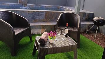 Une terrasse extérieure avec des meubles en osier, une petite table avec une bouteille et des verres, et un élément décoratif aquatique en arrière-plan.
