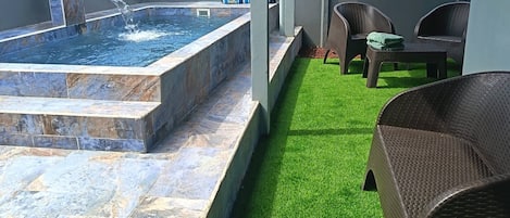 Zona de spa con hidromasaje, césped artificial, mobiliario de mimbre y fuente de agua.