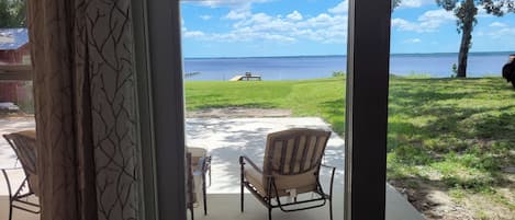 A view through a glass door to a patio with two chairs and a table, overlooking a grassy area and a body of water.