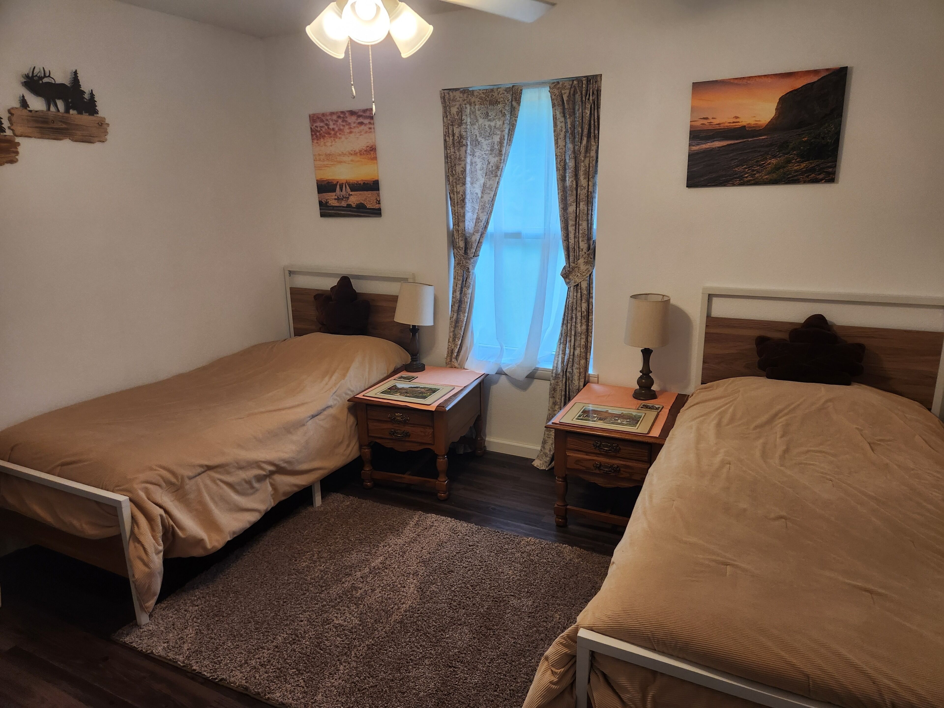 A bedroom with two single beds, a nightstand with a lamp, a window with curtains, and wall art.