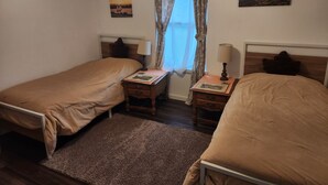 Quarto com duas camas de solteiro, uma mesa de cabeceira com abajur, uma janela com cortinas e decoração na parede.