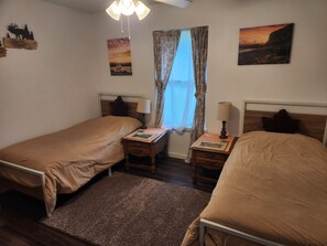 Un dormitorio con dos camas individuales, una mesita de noche con lámpara, una ventana con cortinas y cuadros en la pared.