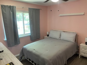 Un dormitorio con cama, mesitas de noche, una ventana con cortinas, un ventilador de techo y una mesilla con lámpara.