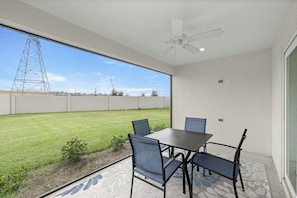 Une terrasse avec une table et des chaises, un ventilateur de plafond et une vue sur une zone herbeuse où se trouve un pylône de ligne électrique.