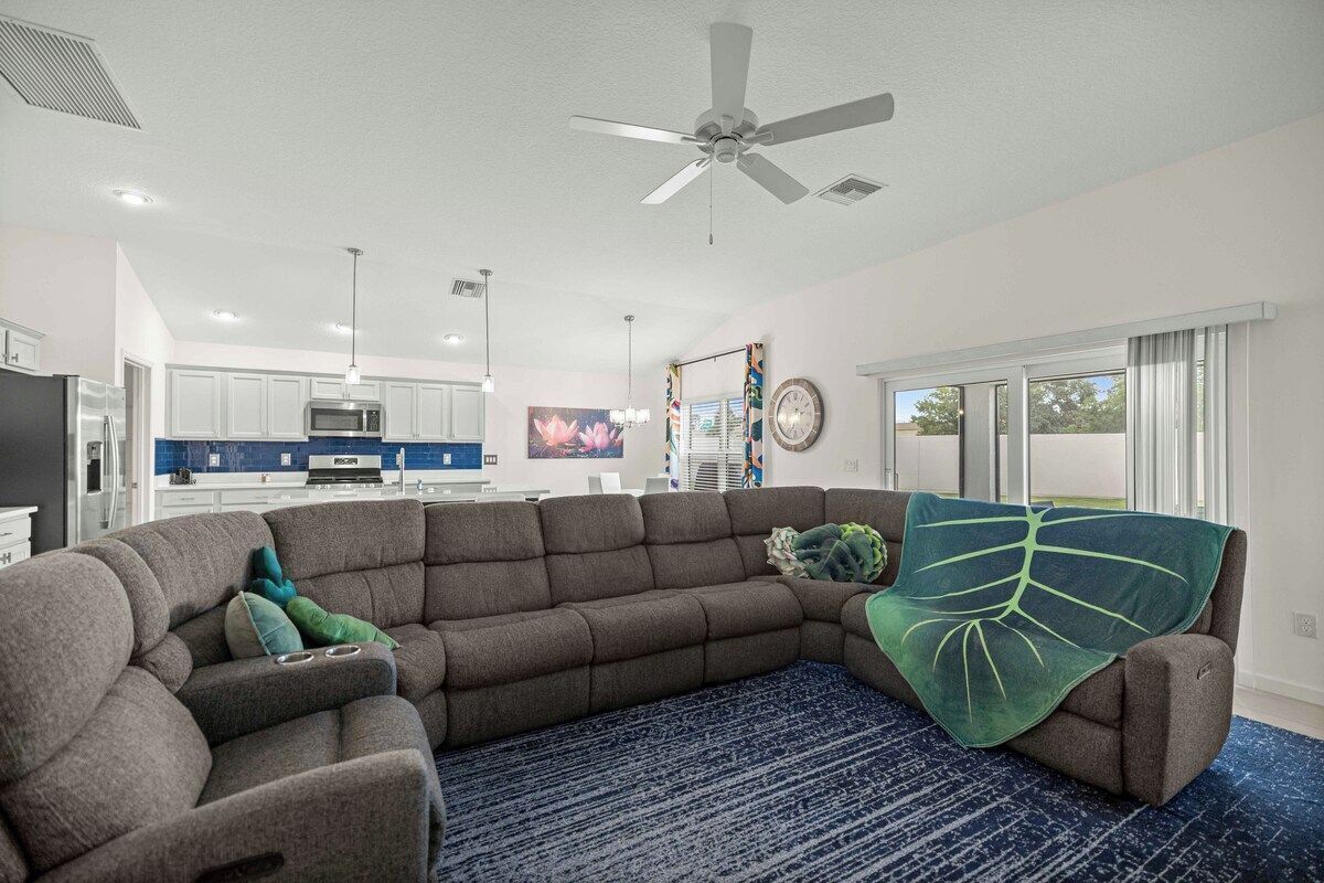 A modern living room with a sectional sofa, a ceiling fan, and a view of the kitchen and dining area.
