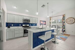 A modern kitchen with white cabinets, a blue backsplash, stainless steel appliances, and a central island with bar stools. - Spacious 3BR · Sleeps 7 · King Bed · Lanai (Okahumpka)