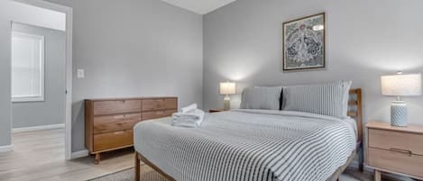 Une chambre à coucher comprenant un lit, des tables de chevet, une commode, une commode en bois, une œuvre d’art encadrée au mur et une porte avec des stores.
