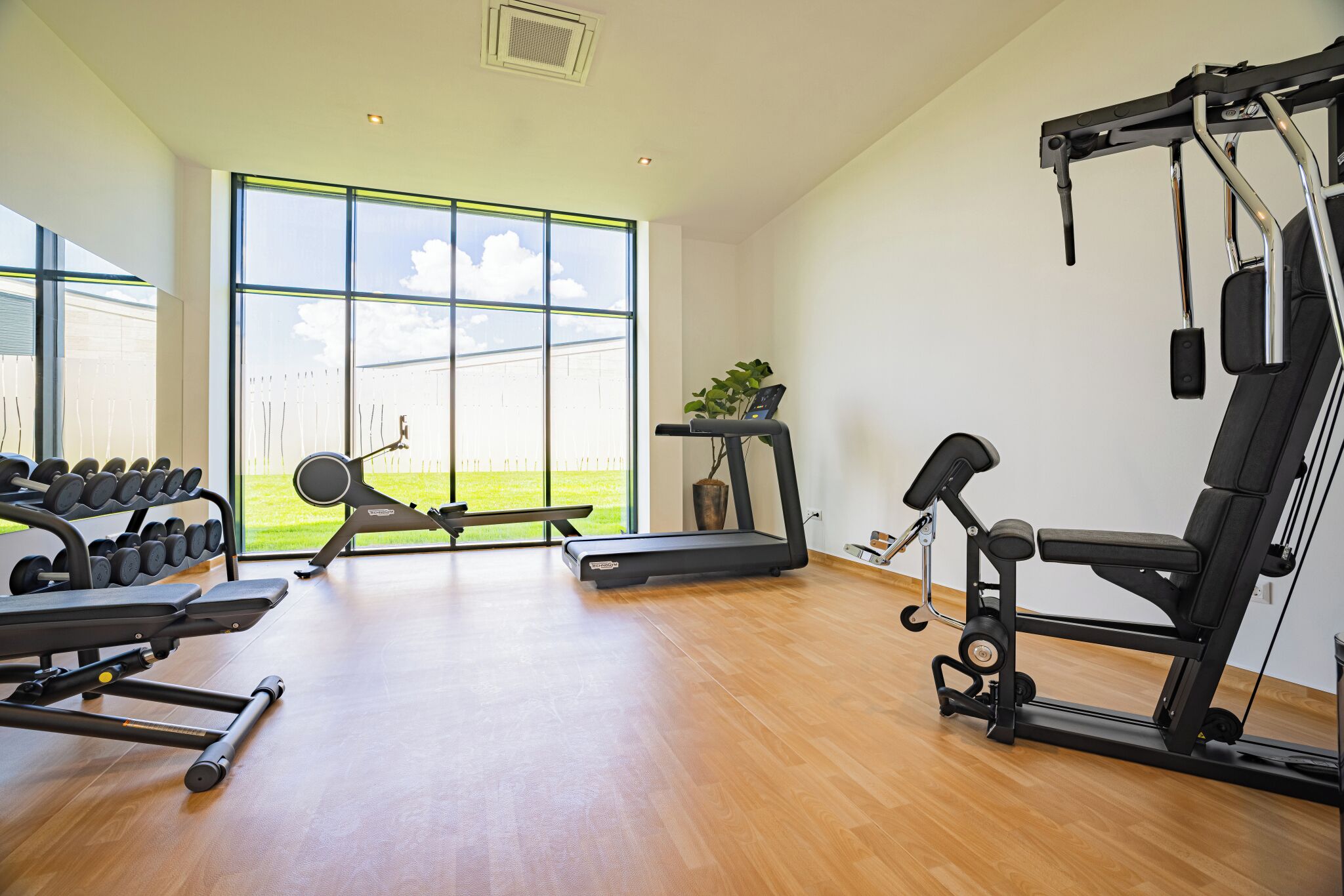 A modern gym with various exercise equipment, large windows, and a wooden floor.