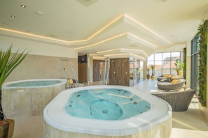 Un área de piscina interior moderna con dos jacuzzis, un espacio de relajación con asientos y vistas a la ciudad a través de amplias ventanas.