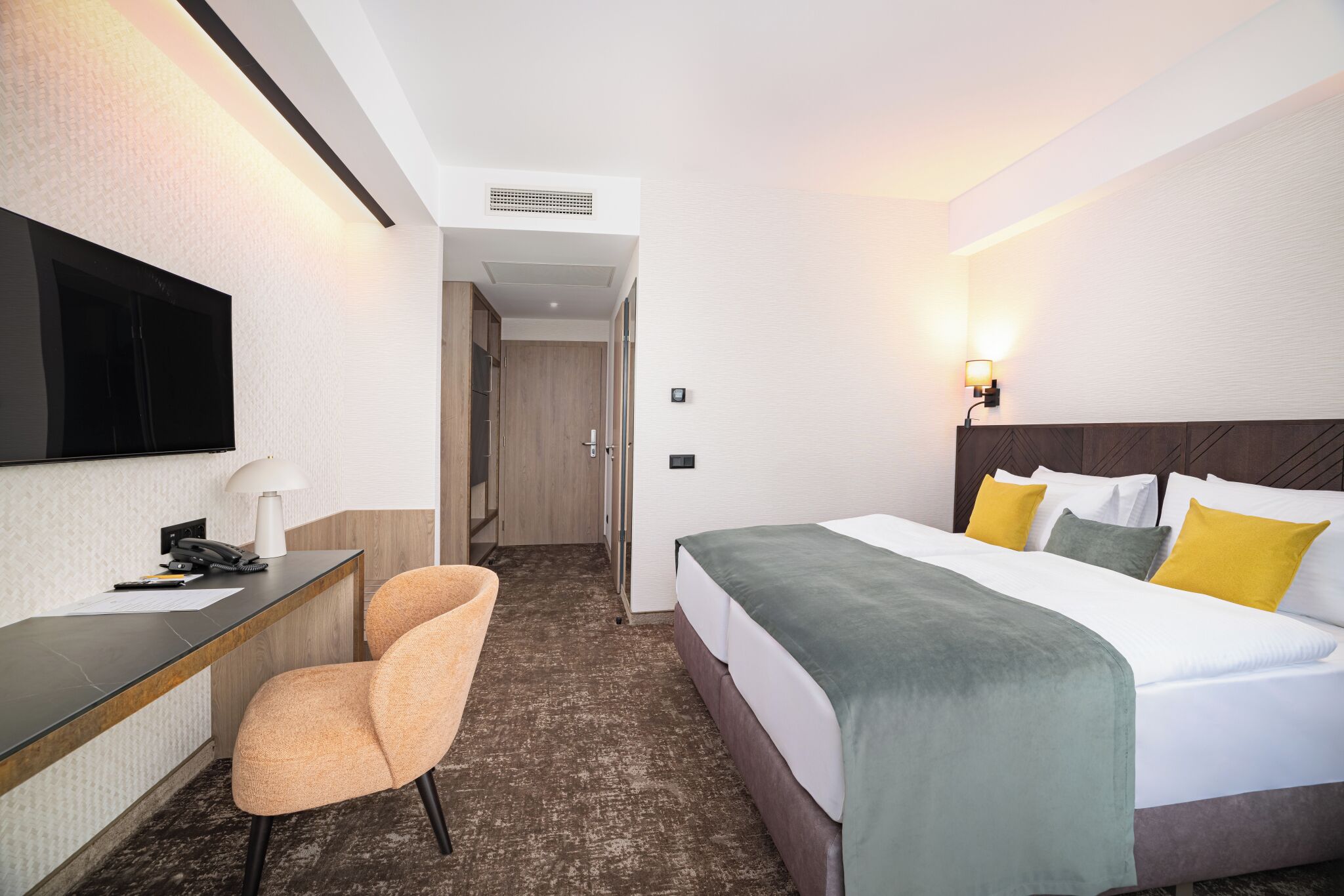 A hotel room with a bed, a desk, a chair, a television, and a wall-mounted light.