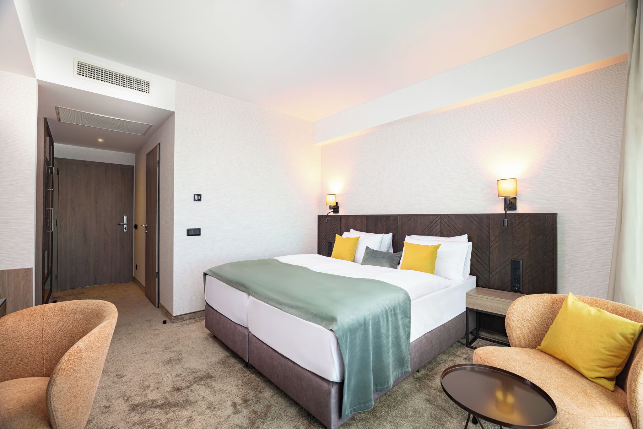 Habitación de hotel con una cama, dos lámparas de noche, un sofá, una mesita y una puerta que da a un pasillo.