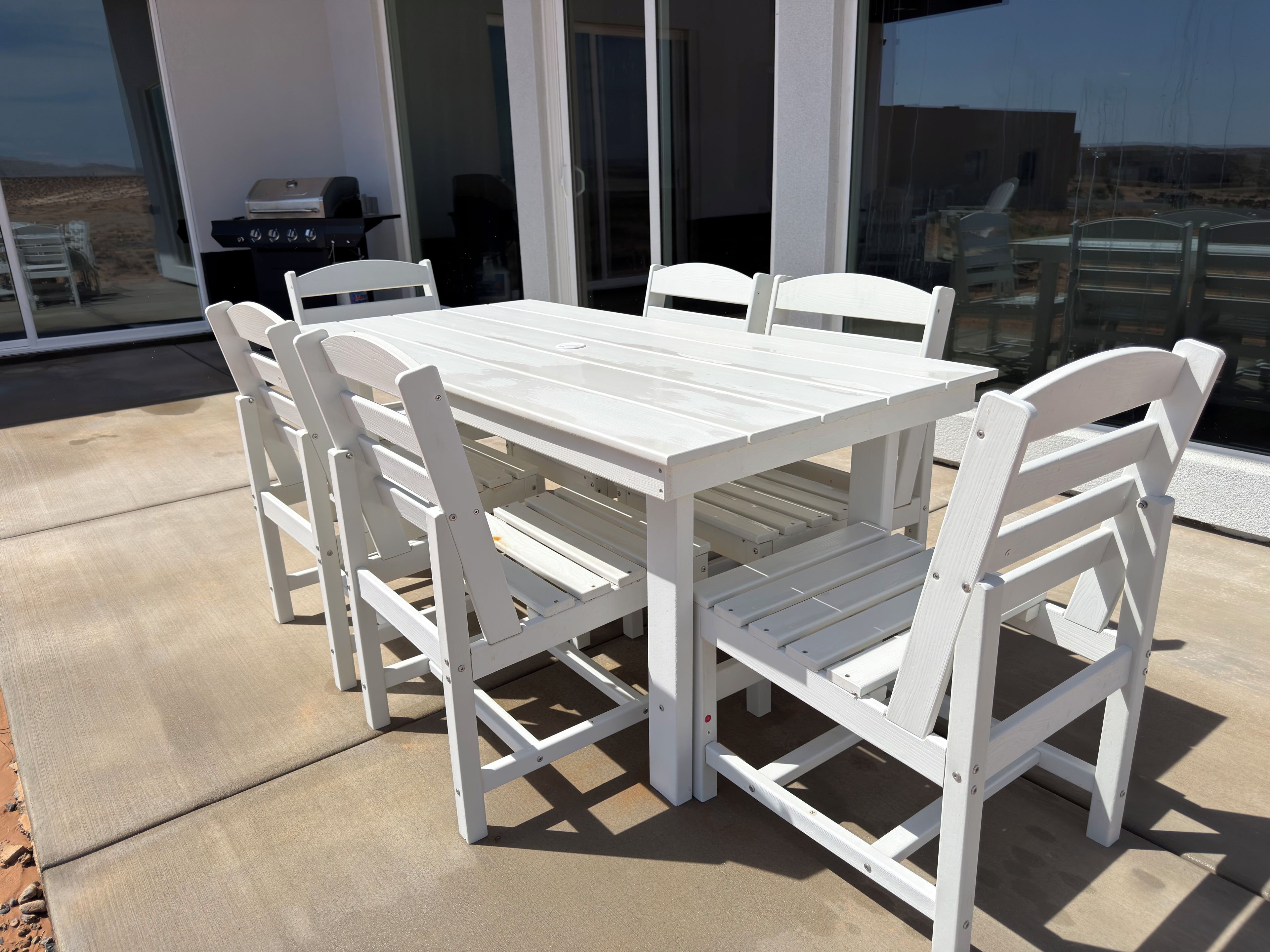 A white outdoor table and chairs set on a concrete patio with a grill and chairs visible in the background.