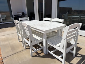 A white outdoor table and chairs set on a concrete patio with a grill and chairs visible in the background.