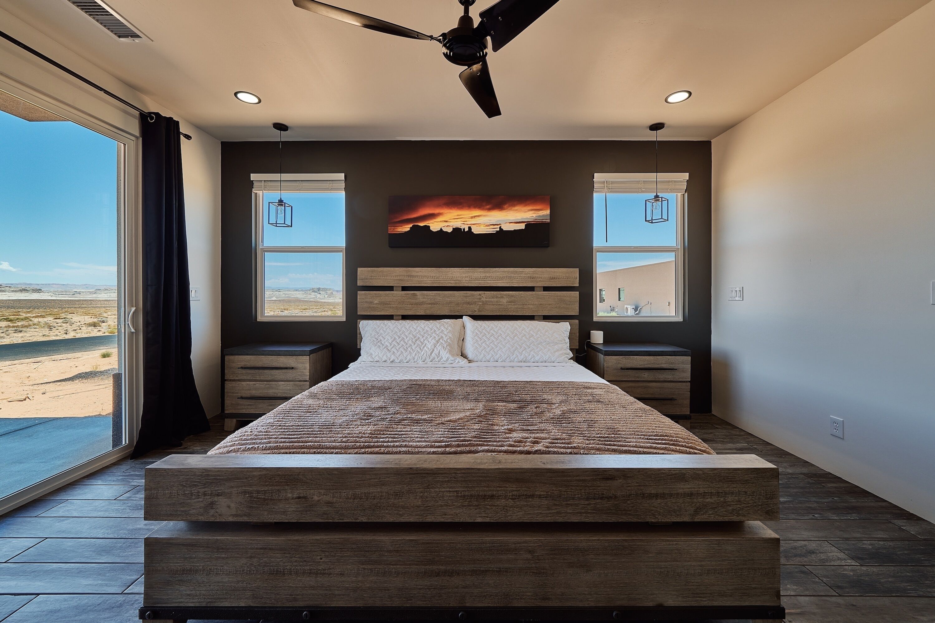 A bedroom with a large bed, a ceiling fan, two bedside tables, a mounted TV, and a view of the desert through the window.