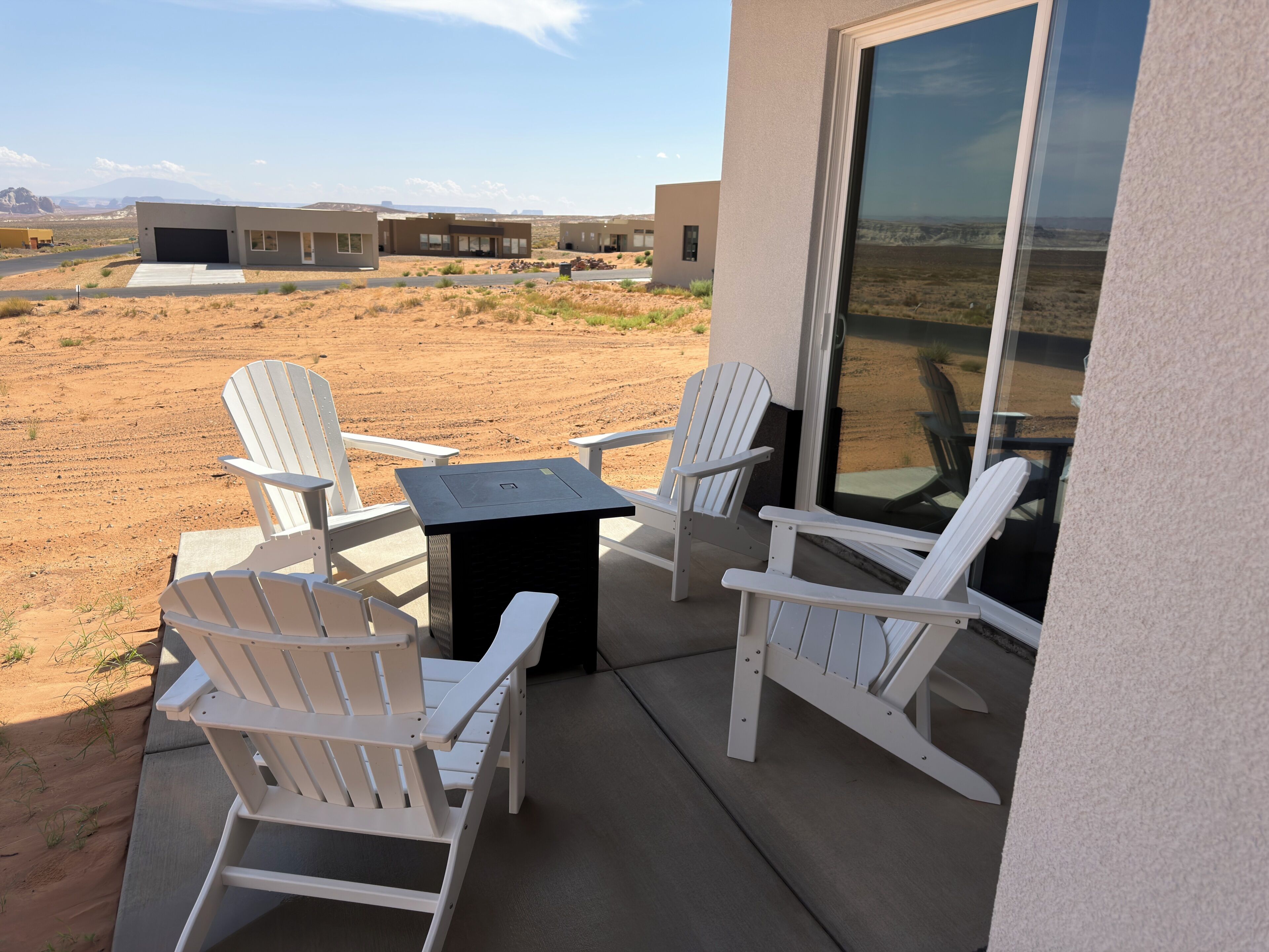 Un patio con poltrone Adirondack bianche e un piccolo tavolo, affacciato su un paesaggio desertico con edifici moderni sullo sfondo.