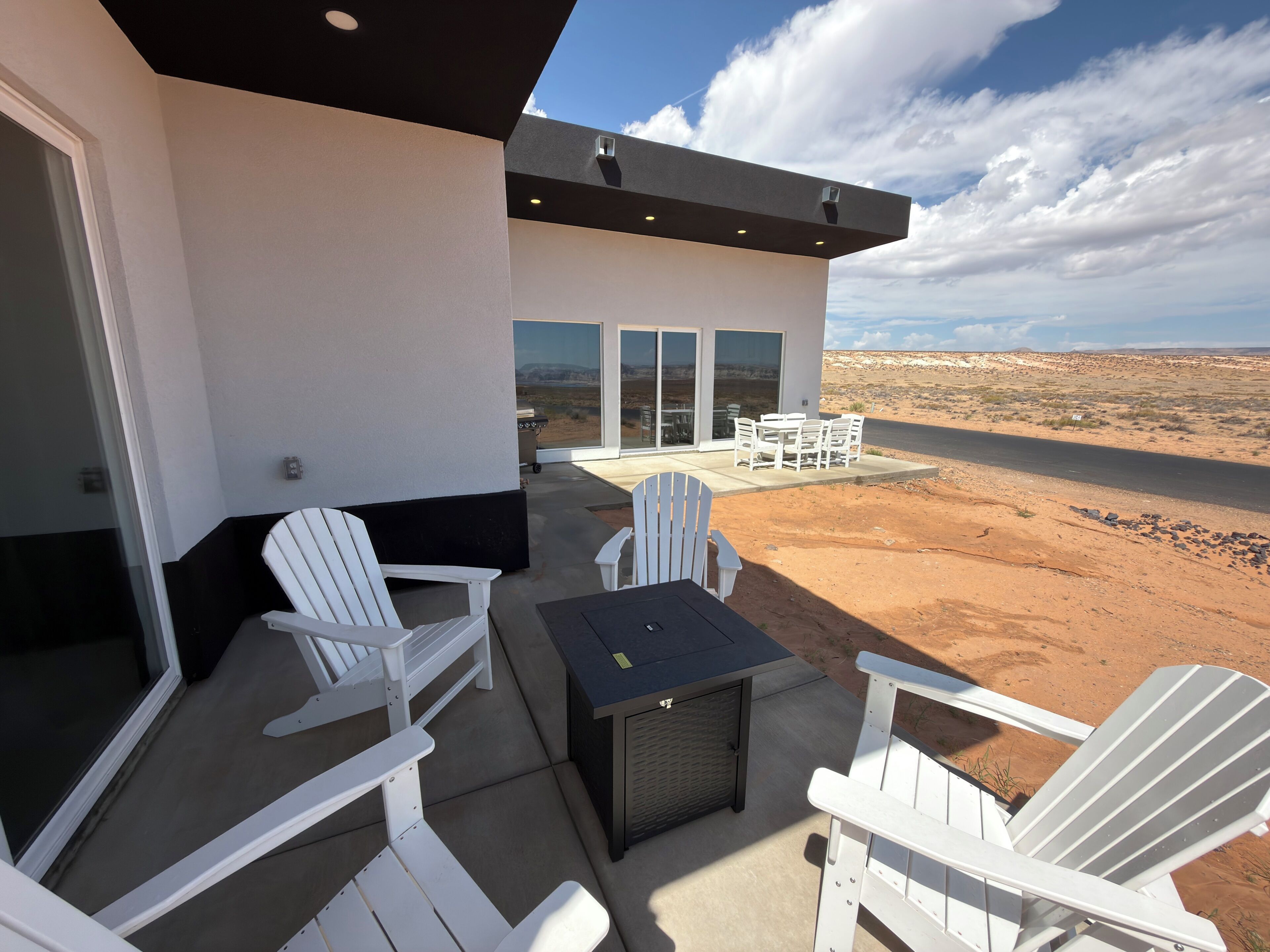 A modern outdoor patio with white Adirondack chairs, a small table, and a glass door leading to a covered area.