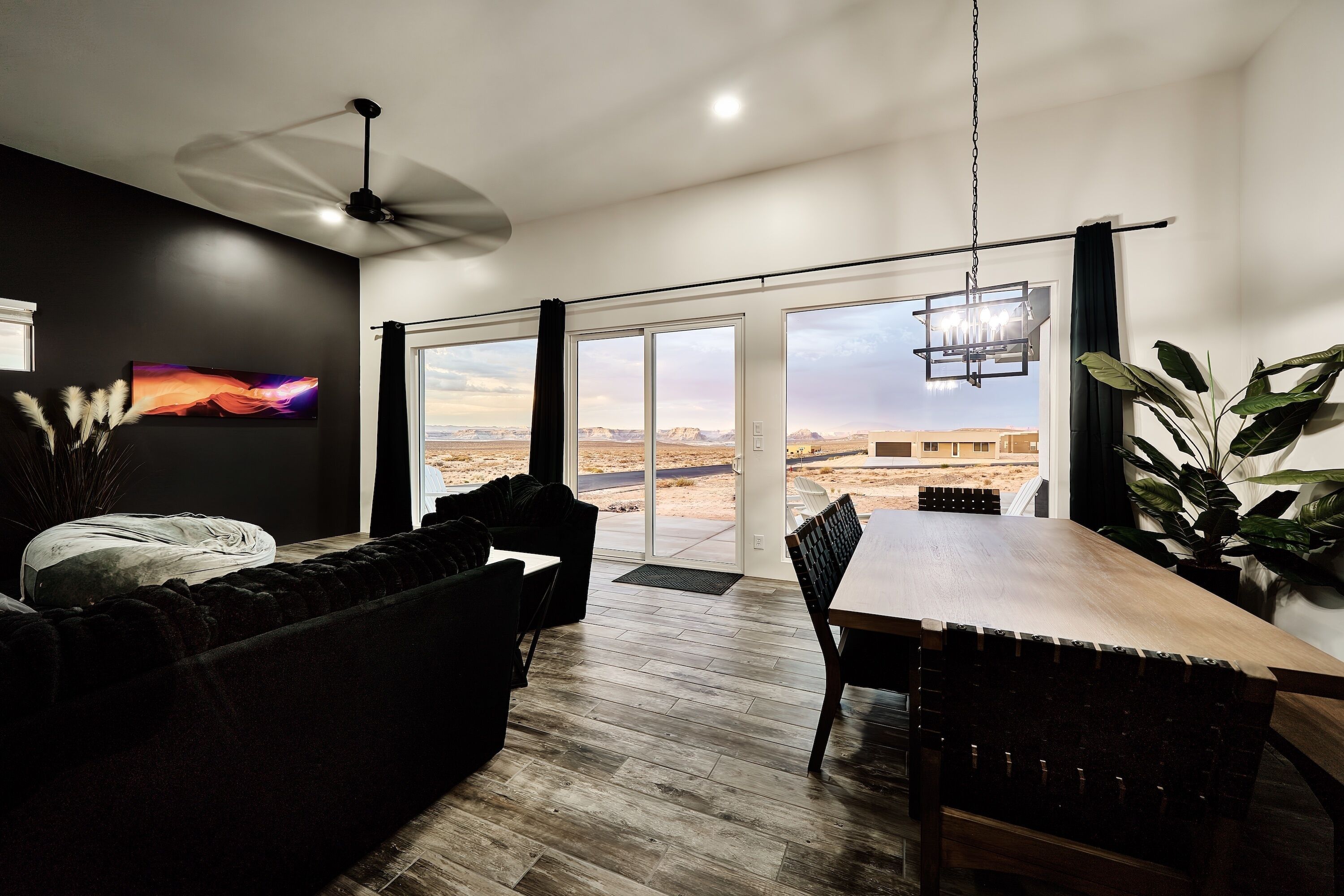 A modern living room with a large dining table, a ceiling fan, and a view of a desert landscape through large sliding glass doors.