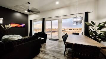 A modern living room with a large dining table, a ceiling fan, and a view of a desert landscape through large sliding glass doors.