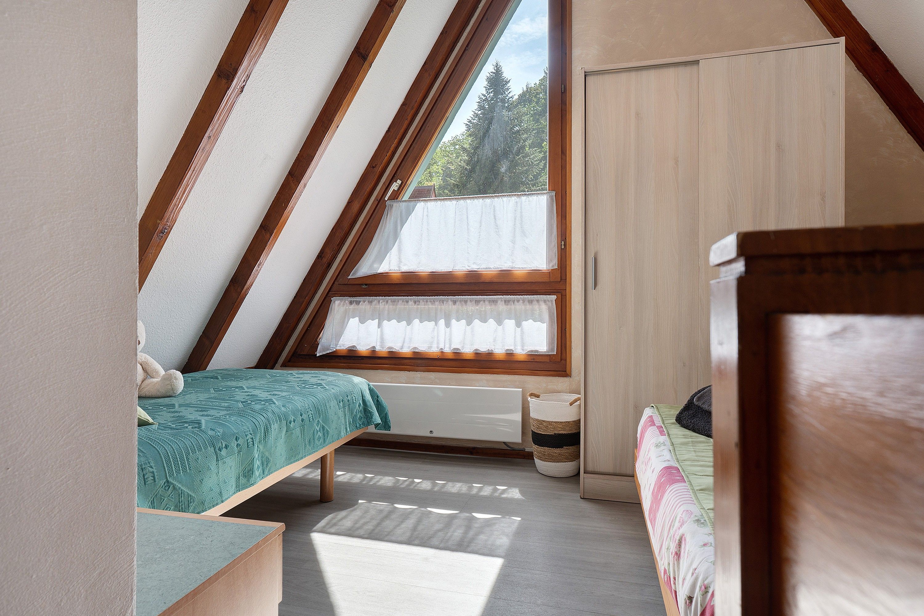 A bedroom with a sloped ceiling, a bed with a green bedspread, a radiator, and a window with a white curtain.