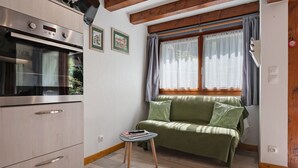 Une cuisine compacte dotée d’un four encastré, d’une petite table et d’un canapé vert placé près d’une fenêtre ornée de rideaux.