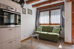 Cocina compacta con horno integrado, una mesita y un sofá verde junto a una ventana con cortinas.