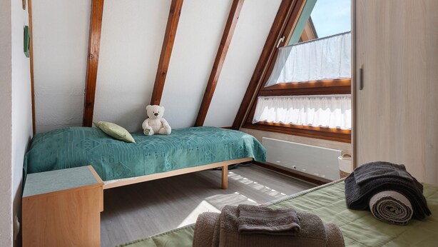 Une petite chambre mansardée, chaleureuse et accueillante, avec un plafond en pente, un lit simple, une fenêtre avec des rideaux et un ours en peluche sur le lit.