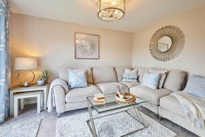 A modern living room with a beige sofa, a glass coffee table, and a decorative mirror. - Host & Stay | Stratton Close (Brotton)