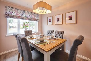 A dining room with a wooden table set for four, upholstered chairs, a window with a floral valance, and three framed pictures on the wall. - Host & Stay | Stratton Close (Brotton)