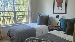 A bedroom with two beds, a window with a view of trees, a framed picture on the wall, and a bedside table with a lamp.