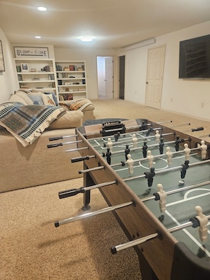 A room with a foosball table, a sofa, a bookshelf, and a sign that reads 'GO JUMP in the LAKE'.