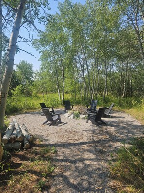 A campsite with four Adirondack chairs arranged around a central fire pit, surrounded by a gravel area with trees in the background.