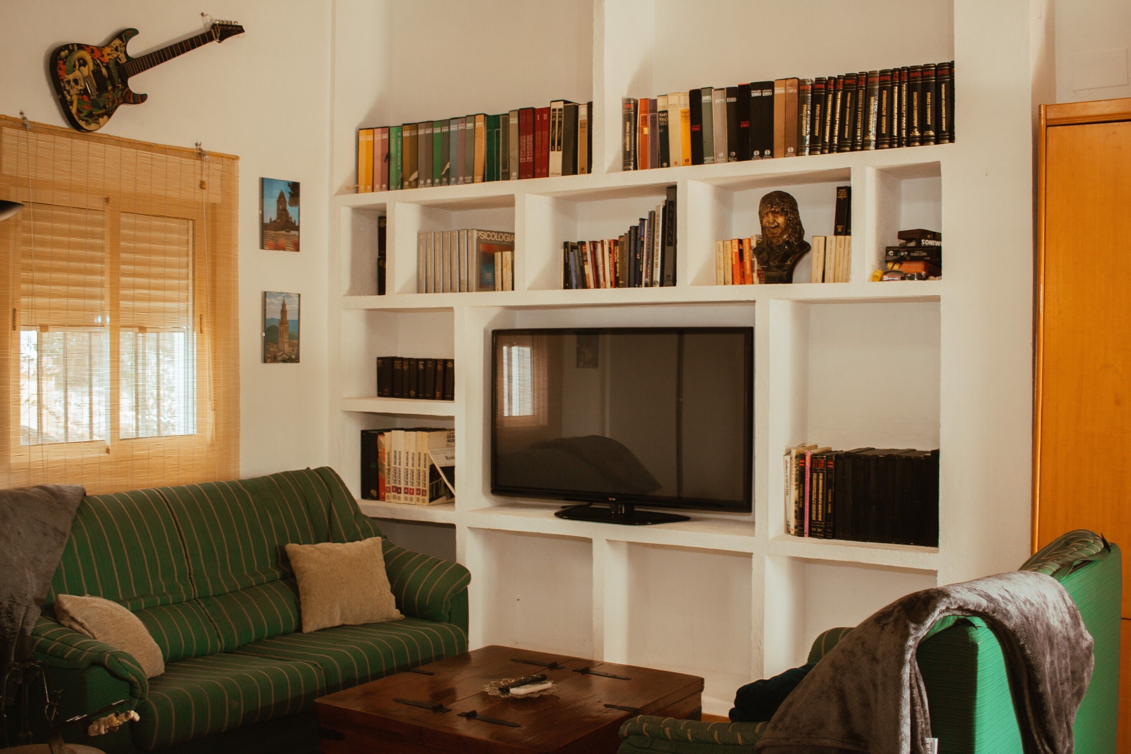 Een woonkamer met een groene bank, een salontafel, een televisie en boekenplanken vol boeken en een buste.