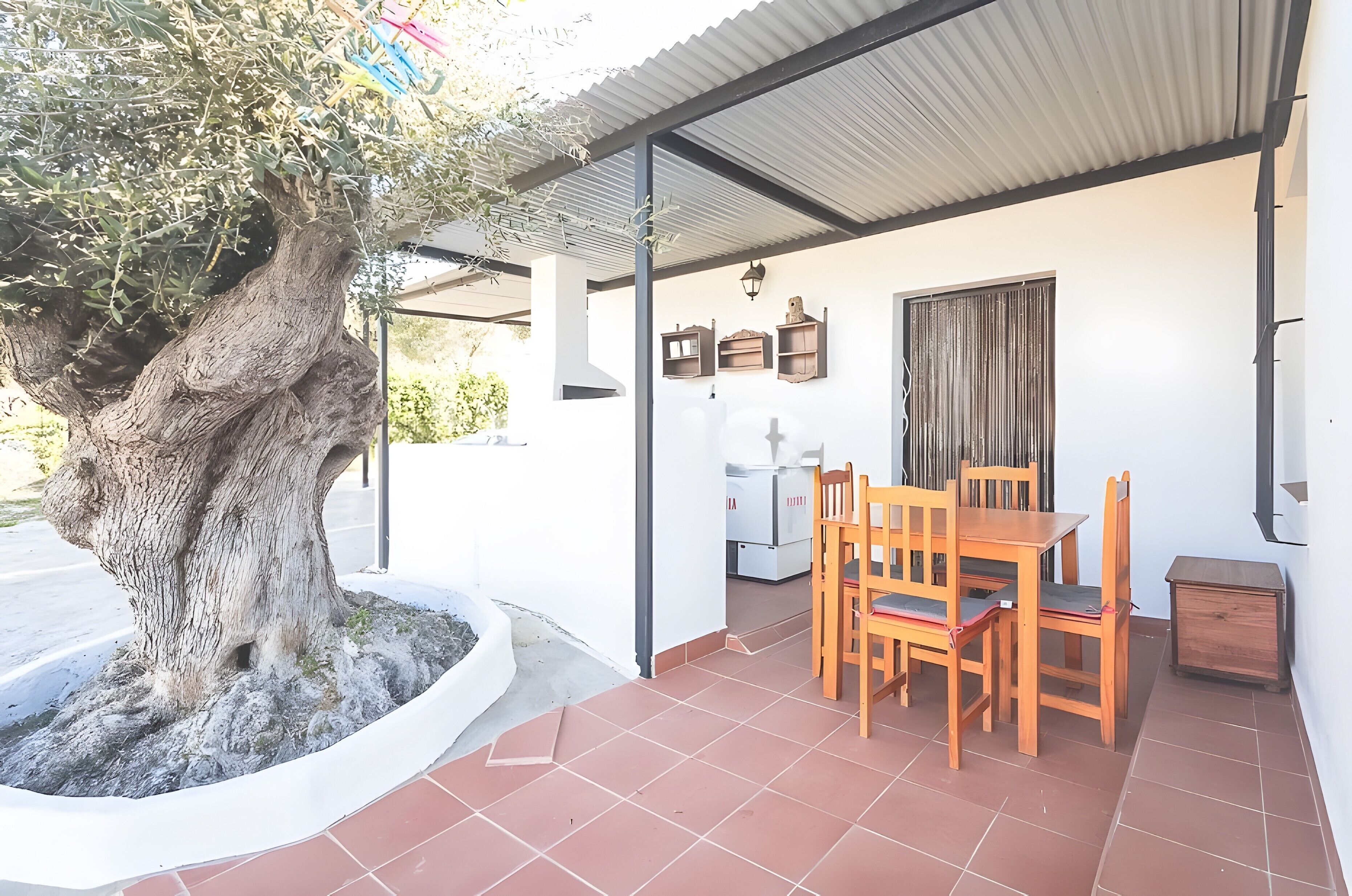 Une terrasse couverte avec une table et des chaises, un grand arbre et un toit en tôle ondulée.
