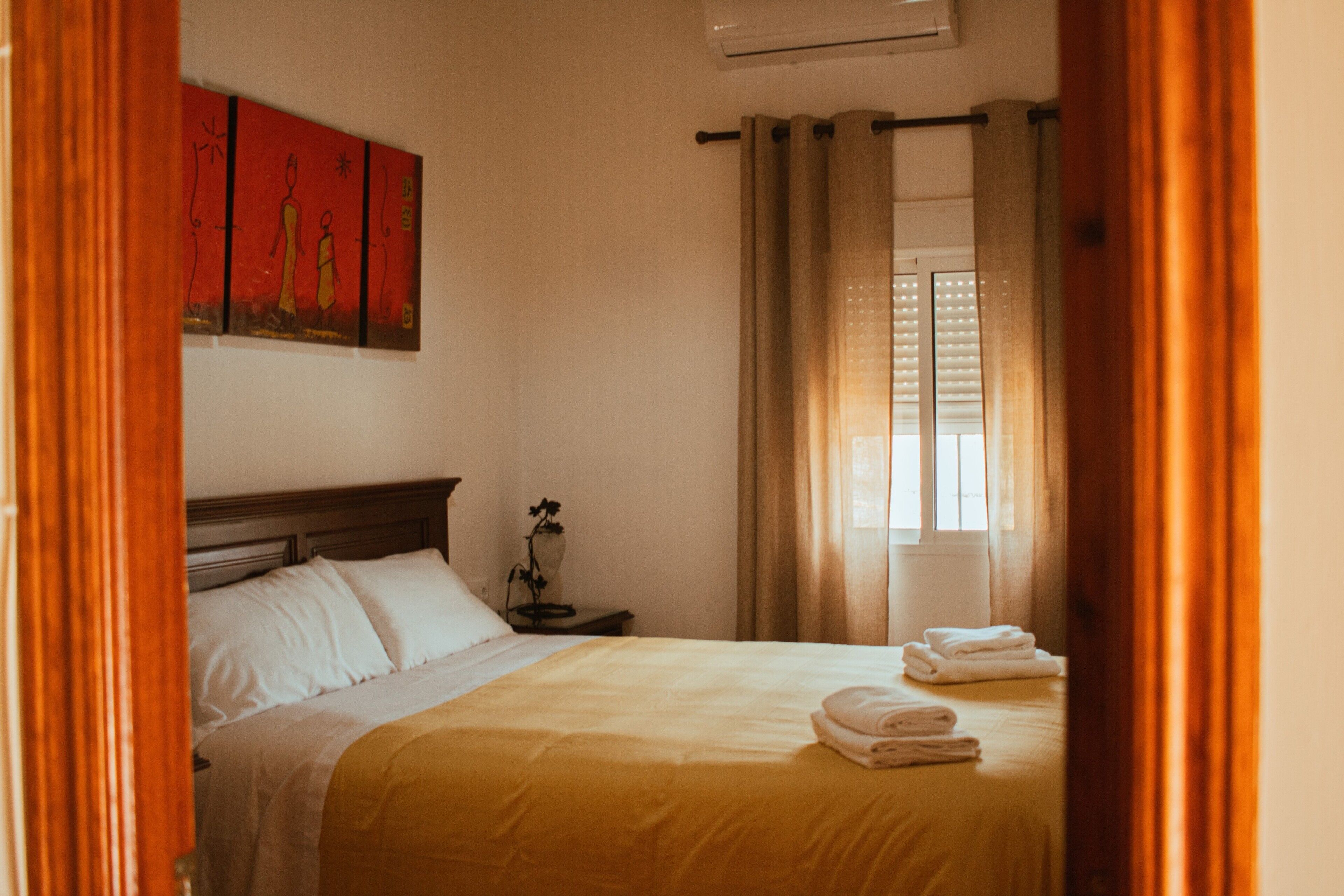 Habitación de hotel con cama, mesita de noche, ventana con cortinas y un cuadro en la pared.