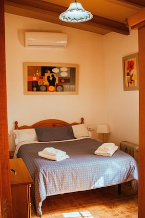 A bedroom with a wooden bed, a bedside table, a lamp, an air conditioning unit, and a painting on the wall.