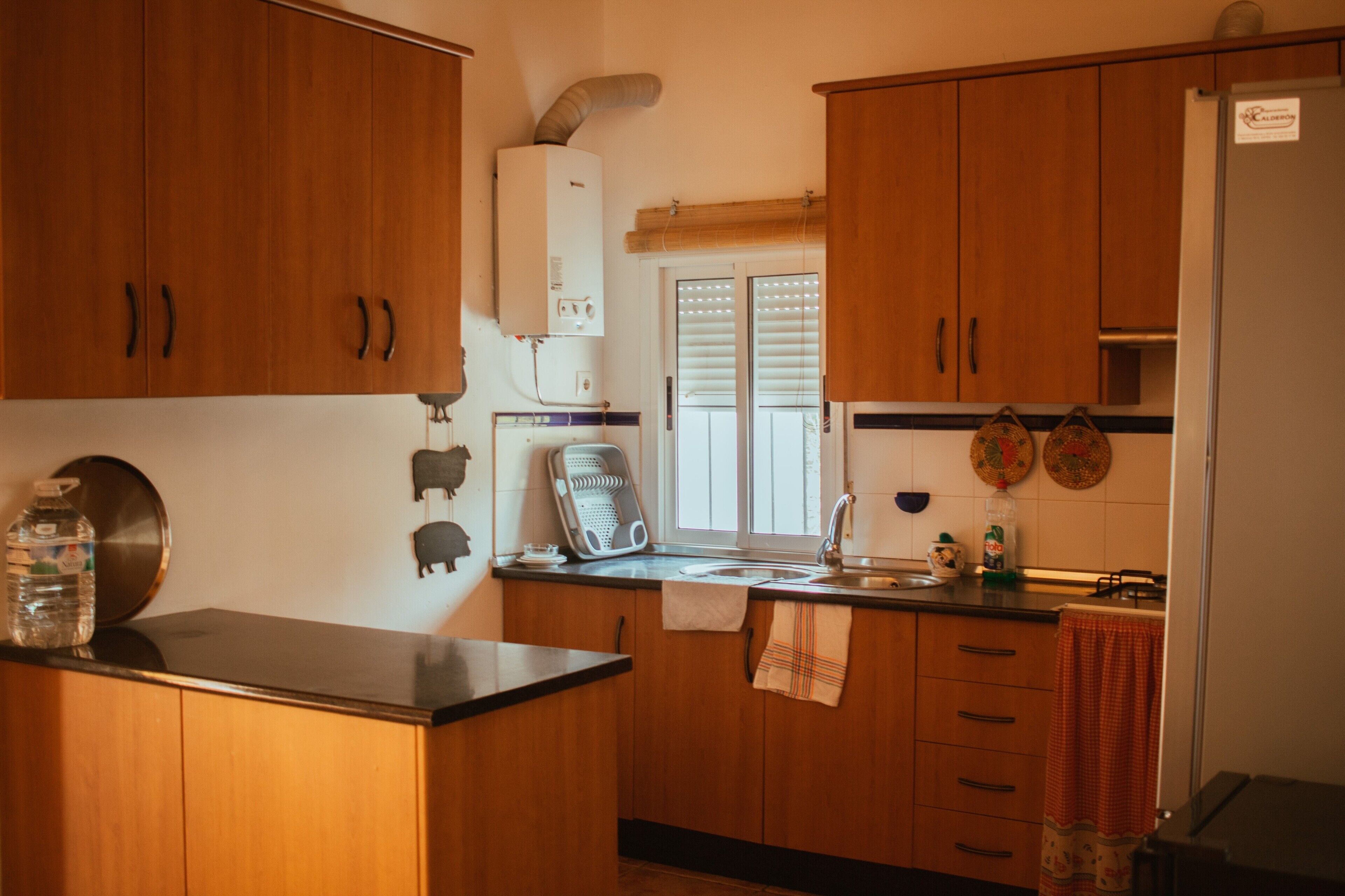 A kitchen with wooden cabinets, a water dispenser, a kettle, and a window with blinds.