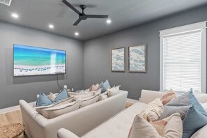Ein modernes Wohnzimmer mit Flachbildfernseher, Deckenventilator und einem großen Fenster mit Jalousien.