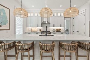 A modern kitchen with white cabinets, a central island with bar stools, and three wicker pendant lights. - Luxury Coastal Retreat in Miramar Beach (Miramar Beach)