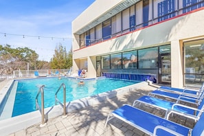 Uma piscina com um terraço solar, espreguiçadeiras e um edifício com grandes janelas.