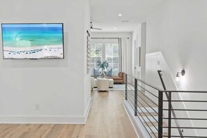 Um interior moderno com uma TV de tela plana na parede, uma escada com corrimão de metal e vista para uma praia pela janela.
