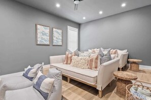 A modern living room with a grey wall, a white sofa, and a matching armchair. There are two framed artworks on the wall, a ceiling fan, and a small round table with a basket. - Luxury Coastal Retreat in Miramar Beach (Miramar Beach)