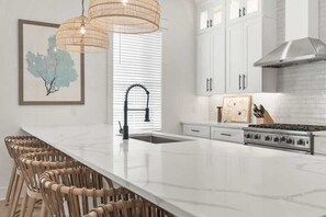 A modern kitchen with white cabinets, a large island with bar stools, a stainless steel range hood, and a framed artwork on the wall. - Luxury Coastal Retreat in Miramar Beach (Miramar Beach)