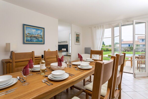 Un coin repas avec une table en bois dressée pour quatre personnes, des assiettes blanches, des verres à vin et des couverts. Des chaises sont disposées autour de la table, et un tableau est accroché au mur.