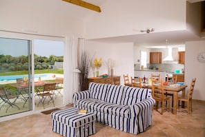 Ein modernes Wohnzimmer mit einer Couch und Hocker, ein Essbereich mit Holzsesseln, eine Küche mit Holzschränken und eine große Schiebeglas-Tür, die zu einem Pool führt.